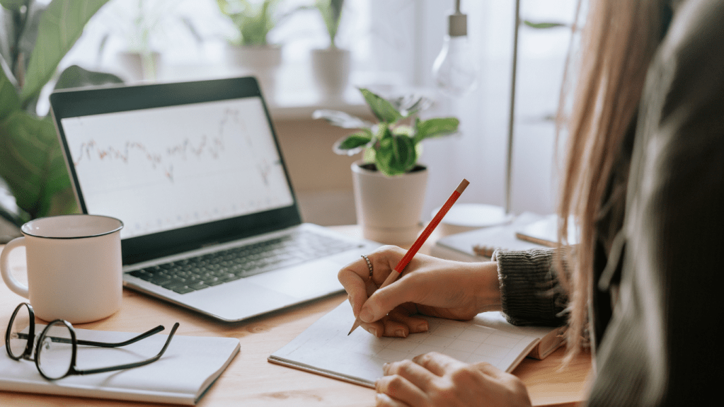 A woman sitting at a desk and writing in her notebook while looking at her laptop.