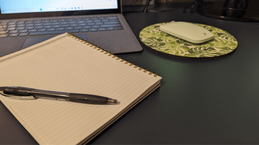 Pen, notebook on a desk with a mouse, mouse page and laptop in the background